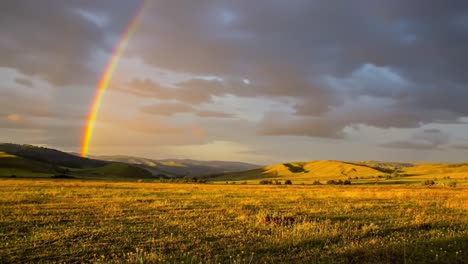a stunning rainbow arches over serene hills, illuminating golden fields and creating a vivid contrast against a dramatic sky filled with clouds