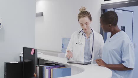 diverse female doctor and nurse using tablet in discussion at hospital reception, in slow motion