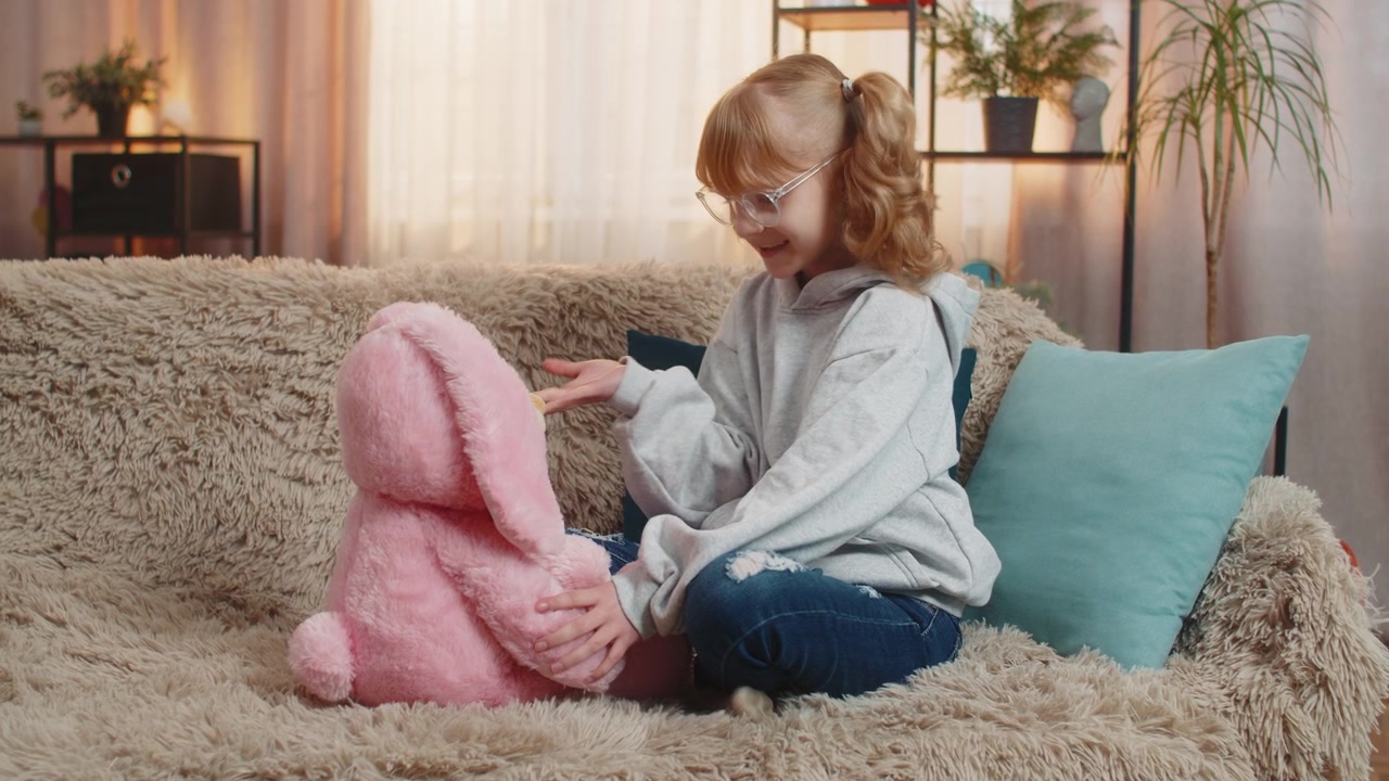 Niña adolescente hablando felizmente con un conejito de peluche en el sofá de casa, simulando un juego divertido e imaginativo