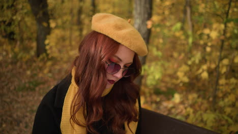 lady in yellow beret, sunglasses, and red wavy hair looking at canvas, tilting head from right to left, immersed in artistic thought, seated on wooden bench surrounded by golden autumn foliage