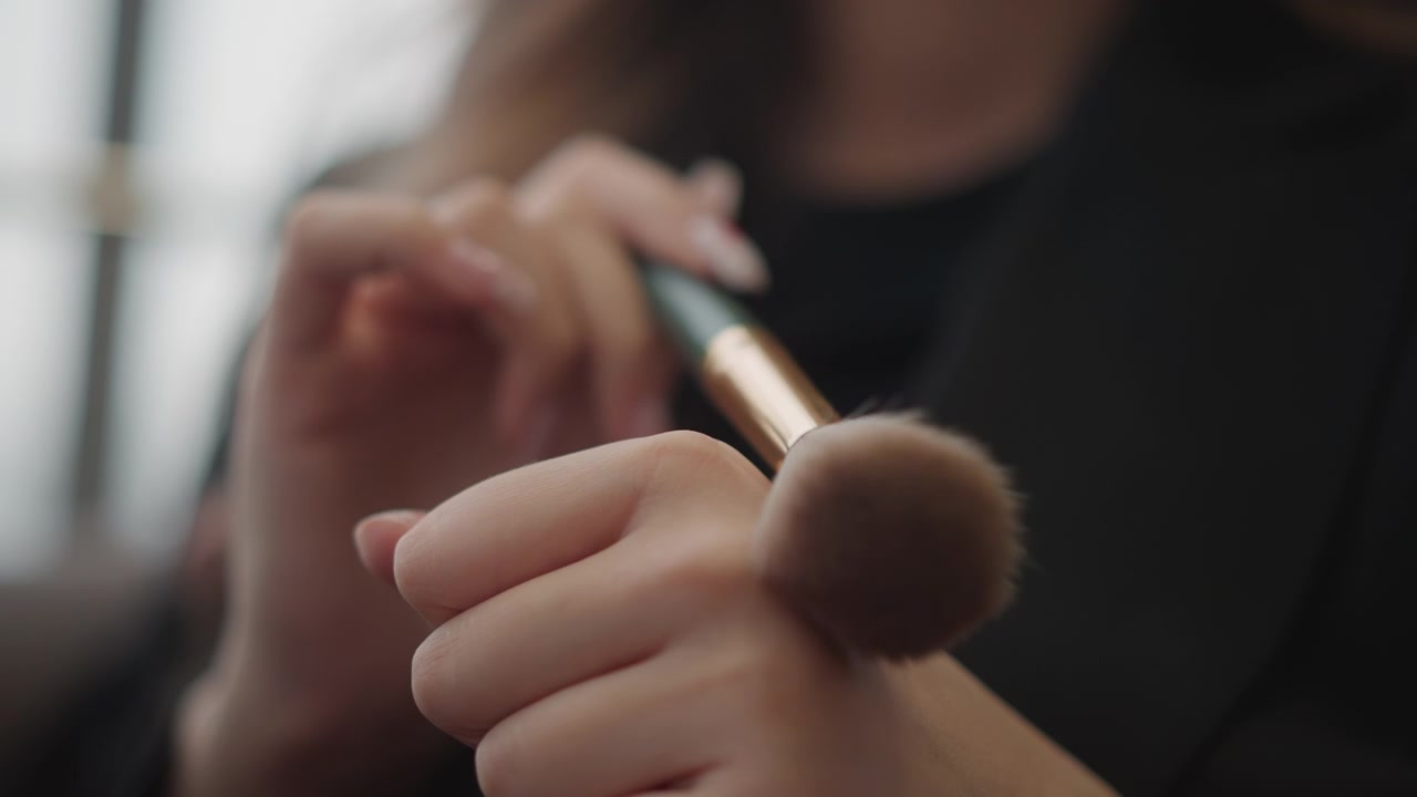 Tight vertical close-up shows hands holding slender brush, sweeping fluffy brush over skin, and guiding small manicure tool, with soft window light, subtle blur, calm beauty routine