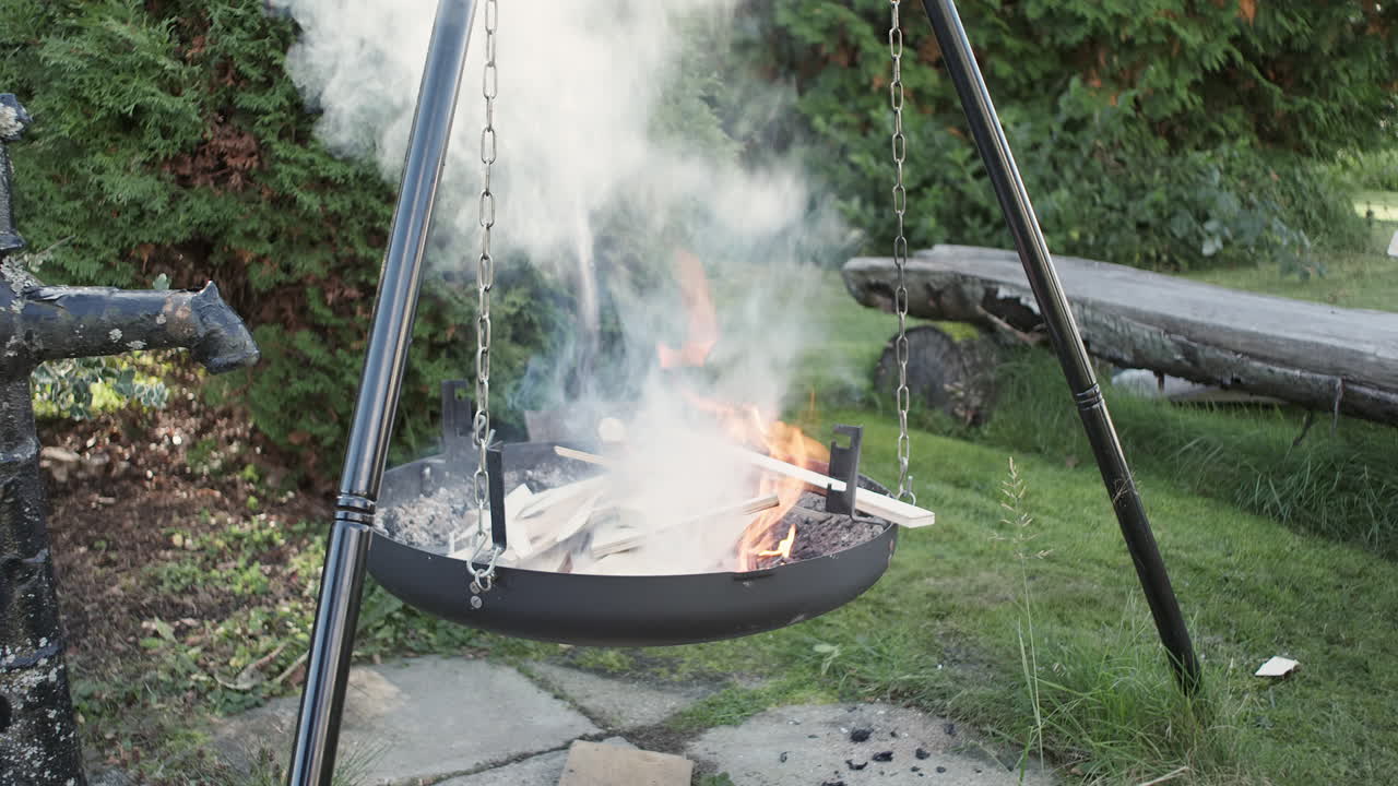 A hanging fire pit with burning wood and smoke rising, placed in a grassy garden area surrounded by trees.