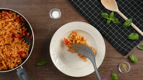 top down shot of kitchen utensil, spatula putting creamy fusilli pasta with tomato sauce on deep plate placed on wooden table decorated with other spatula, black cloth and fresh basil leaves