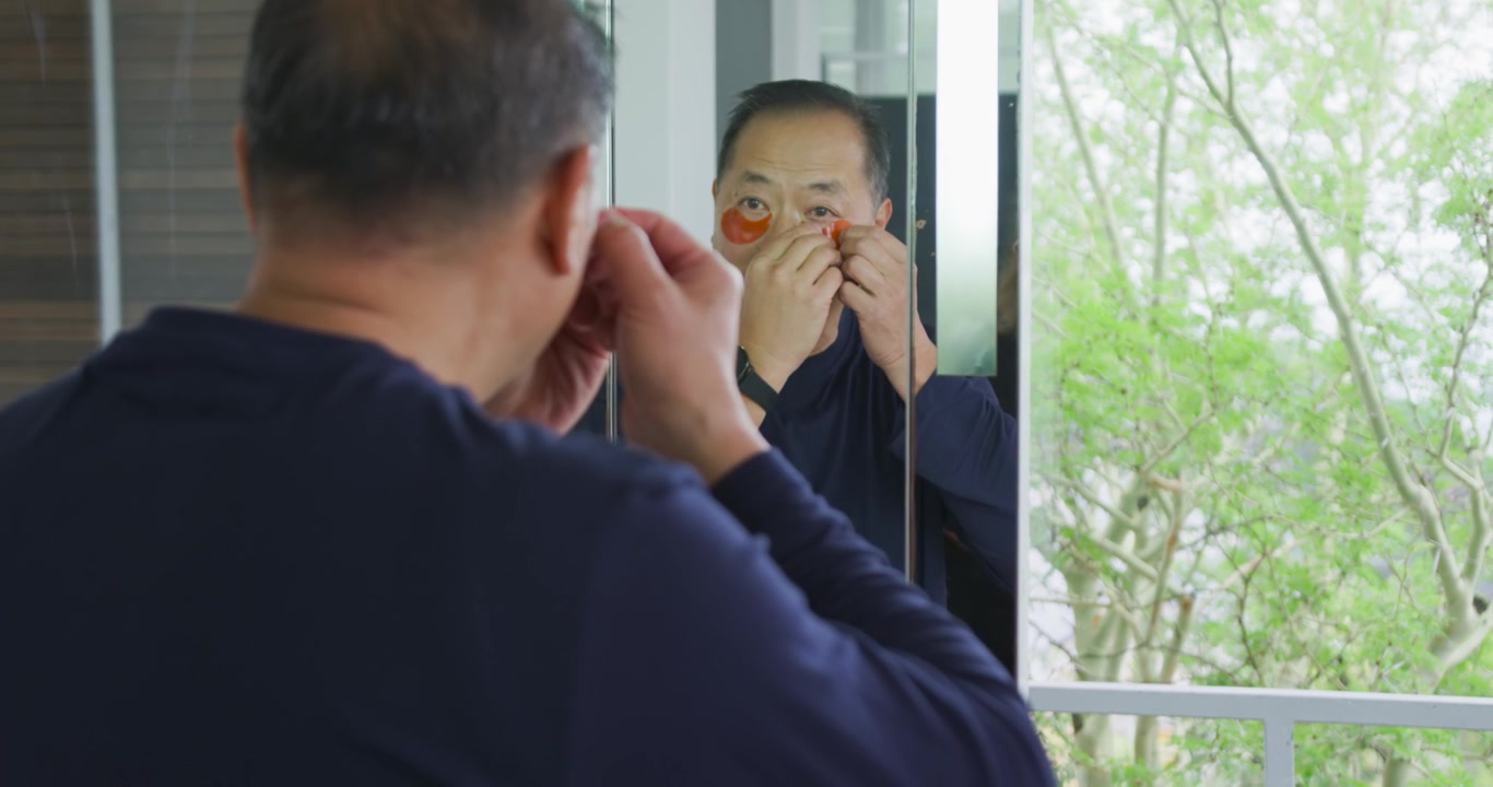 Anziano uomo asiatico che si guarda allo specchio, applicando patch idrogel arancioni sotto gli occhi alla toeletta per la cura della pelle.