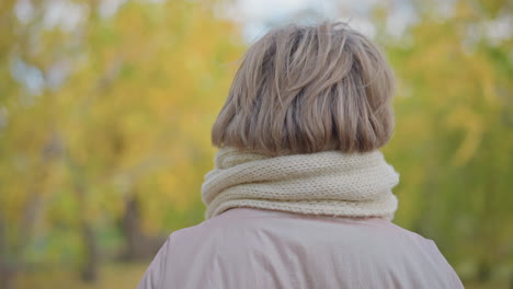 back view of woman wearing pale pink jacket and thick white knitted scarf while adjusting muffler during calm autumn stroll, surrounded by blurred golden and green foliage