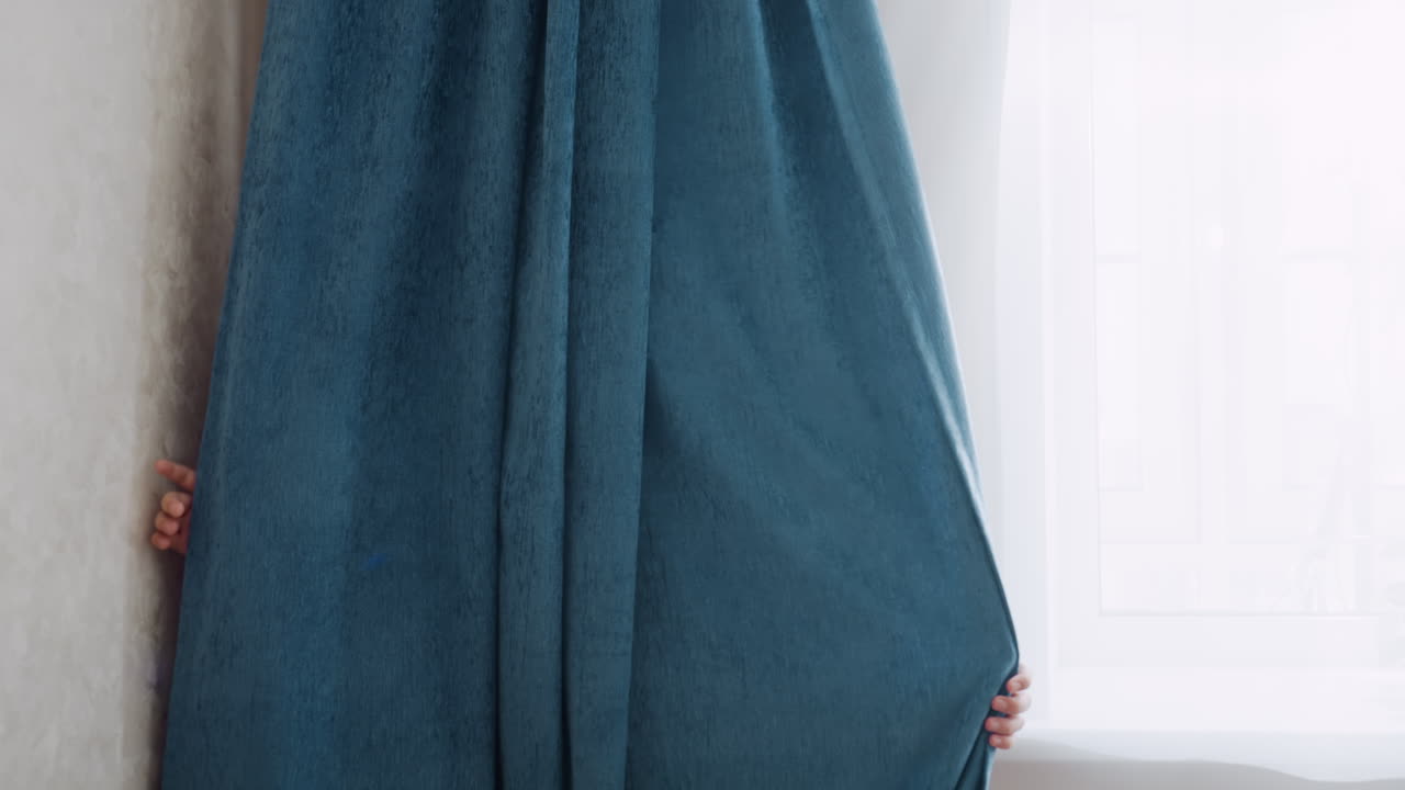 Boy in light green shirt hides behind curtain in bright sunlit room with smile on face during playful hide and seek moment while enjoying fun time indoors with soft daylight and relaxed mood
