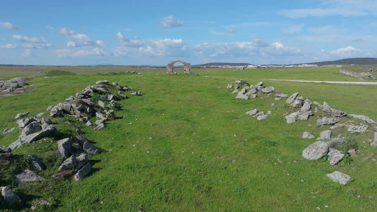Frontal drone flight over pastures and two lines of ruined pigsty structures. The striking arch of Puerta de Arenas remains centered as the key element throughout the composition.