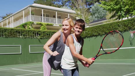 madre caucásica enseñando a su hijo a jugar al tenis en la cancha de tenis en un brillante día soleado