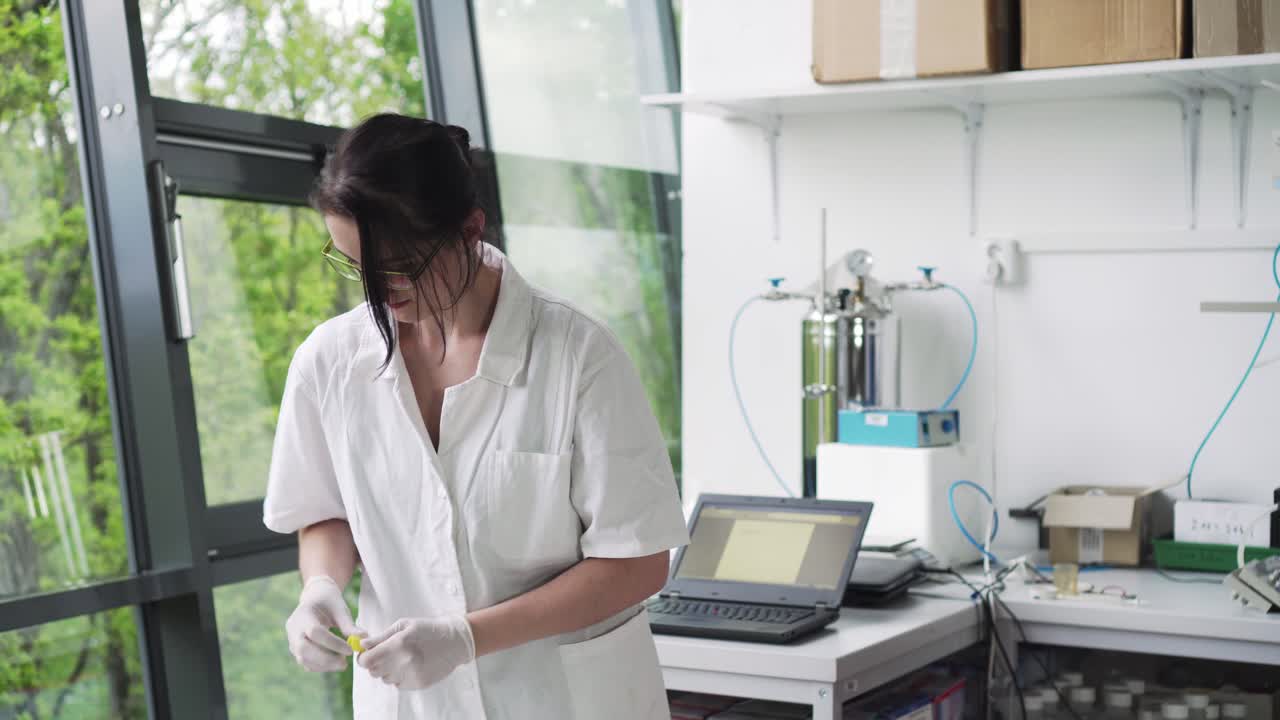 Female researcher using laptop in laboratory and marking test tube, front middle view