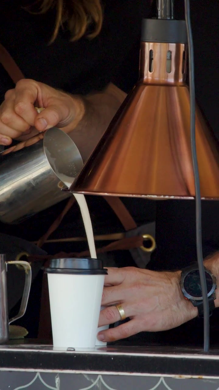 Barista gießt aufgeschäumte Milch in einen Coffee-to-go-Becher unter warmer Beleuchtung, Hände sichtbar, Café-Umgebung, Nahaufnahme.
