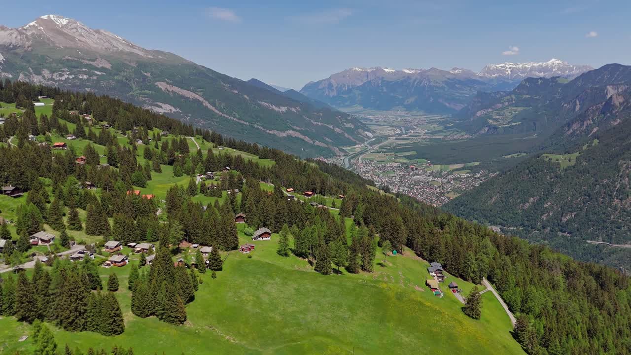 Idyllic small swiss village on hilltop of Alps. Aerial wide shot. Sunny day in summer. Snowy peak of alp mountains in distance. Green uphill landscape.