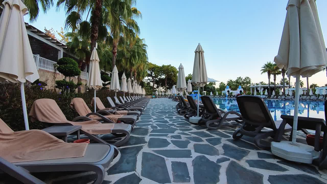 Bir tatil köyü havuzunun kenarında, palmiye ağaçları ve taş döşemelerle çevrili bir sıra şezlong ve şemsiye, rahatlatıcı bir tatil atmosferi yaratıyor. Seyahat, boş zaman ve lüks tatil köyleriyle ilgili ideal temalar.