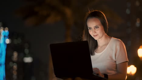 A-young-female-student-with-a-laptop-at-night-in-the-city-looking-at-the-computer-screen-and-typing-with-his-hands-on-the-keyboard.-Remote-work-on-the-Internet.-The-student-does-the-work.-Night-life-in-the-city