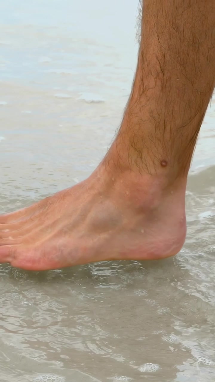 Video verticale: Goccioline che cadono, gamba maschile scalza che entra nella risacca bassa per stare in piedi, neo sulla caviglia.