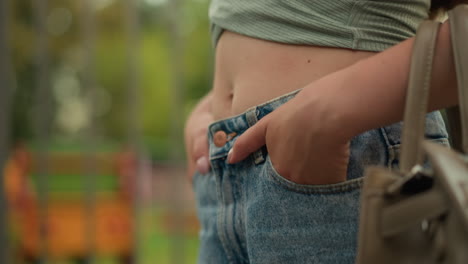 close-up of young woman walking with hands in pockets, exposing midriff, wearing green cropped top and high-waisted jeans, beige handbag on arm, with blur background