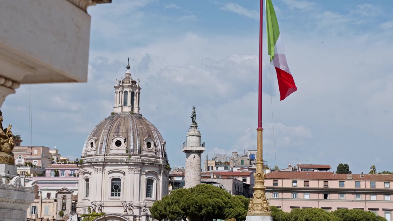 Bandiera tricolore nazionale sventola accanto a una storica cupola di chiesa e alla Colonna Traiana sopra lo skyline del centro città.