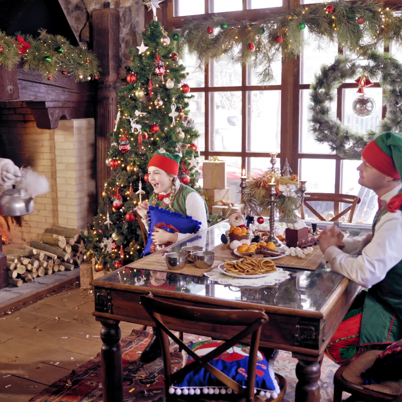 Elves sitting at the table on beautifully decorated background for Christmas. Santa Claus with hot kettle coming to the table.