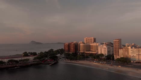 Aerial-panning-down-movement-at-golden-hour-at-the-Copacabana-fort-in-Rio-de-Janeiro-with-the-ocean-waves-coming-in-on-the-beach,-sunlight-hitting-the-buildings-and-islands-in-the-background
