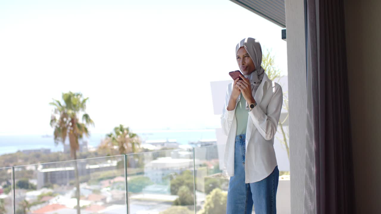 Donna adulta con foulard in testa in piedi sul balcone dopo una notifica di messaggio, che scorre un telefono rosa, con spazio per il testo.