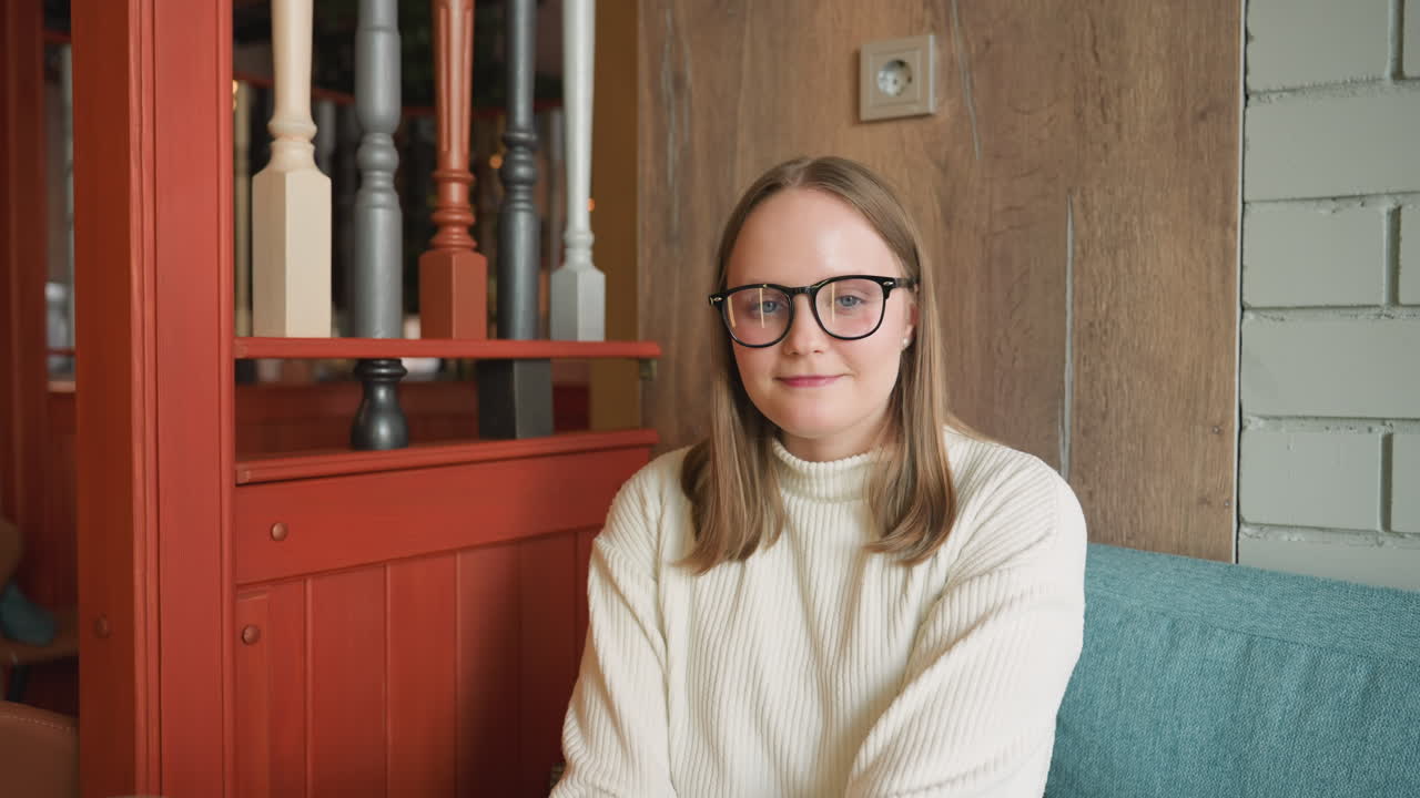 Confident woman in glasses and white ribbed sweater sitting on blue sofa smiling warmly while looking directly at camera, cozy indoor setting with wooden wall and red decorative frame in background