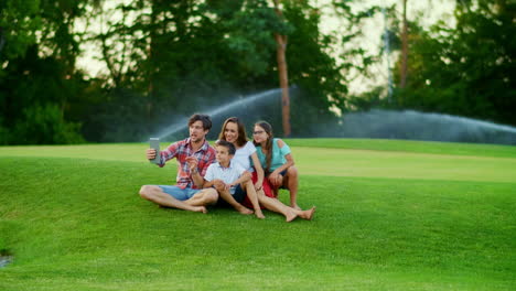 Family-sitting-on-green-in-meadow.-Parents-and-kids-using-digital-tablet-in-park
