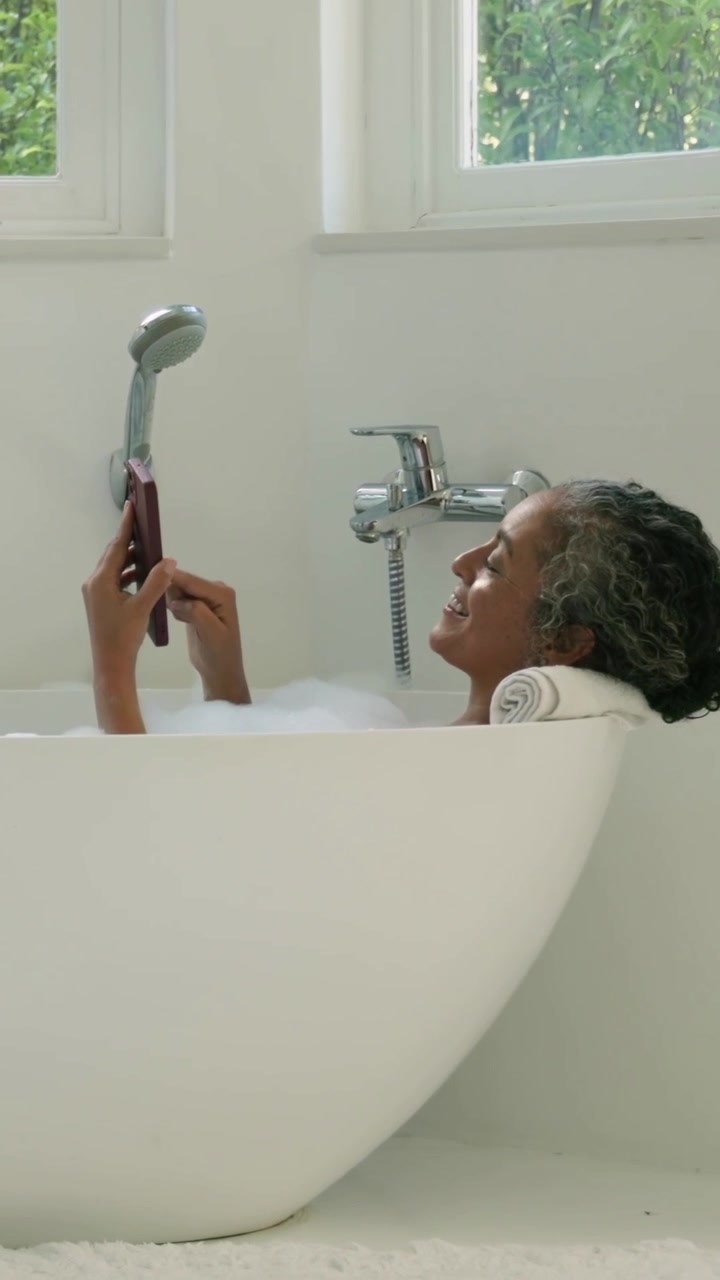 Vertical video: Reclining woman reading, tapping phone and lifting shower in home bath, copy space