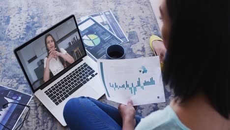 Mixed-race-businesswoman-sitting-on-floor-using-laptop-having-video-call-with-female-colleague