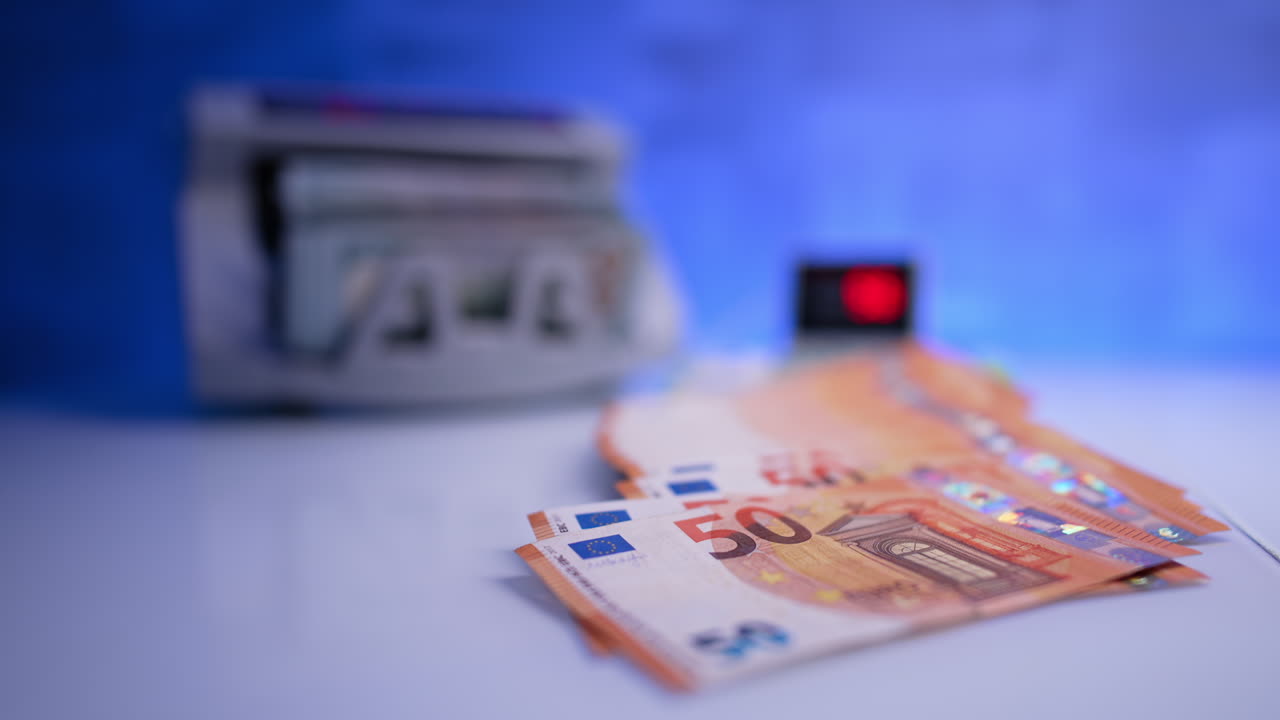 Fifty euro banknotes lie on the desk. Money counting machine works at blurred backdrop and counted cash stack is put at foreground. Close up.
