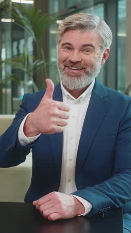 happy mature caucasian businessman in suit looking at camera showing thumb up in modern office lobby