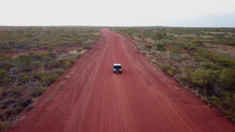 Aerial:-Drone-shot-tracking-a-solo-vehicle-driving-down-a-red-dirt-track-towards-the-horizon-during-early-evening,-just-outside-of-Alice-Springs,-Australia
