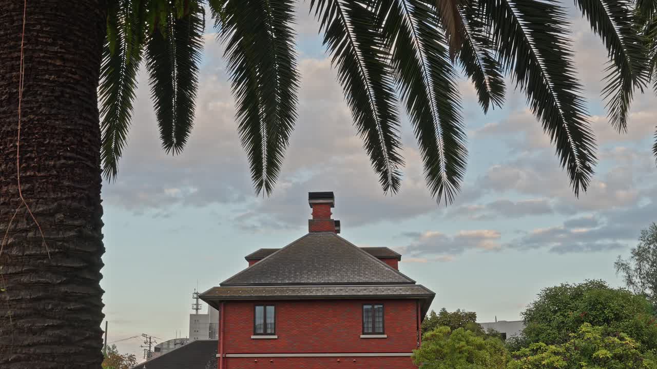 A distinct red brick building with tall chimneys framed by the dramatic leaves of a large palm tree against a cloudy sky