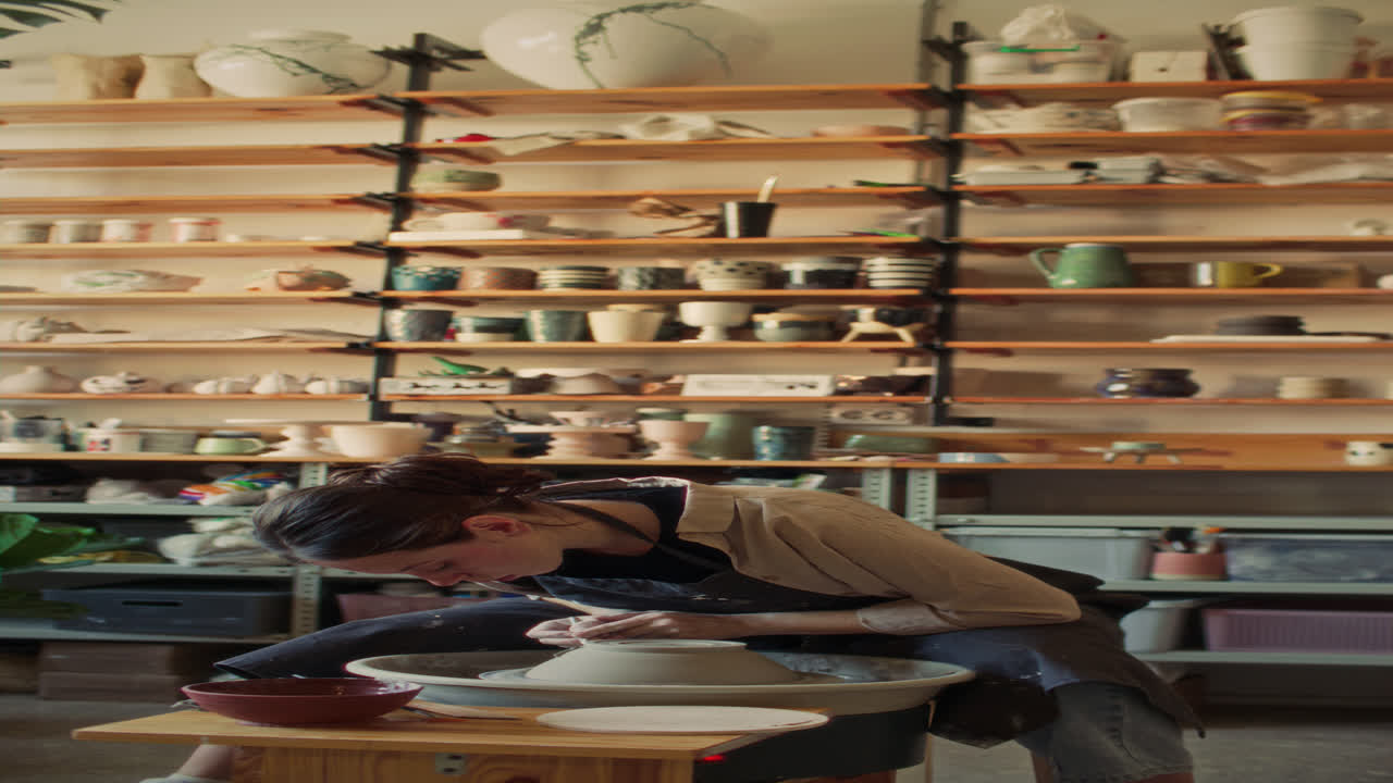 Female Artisan Trimming Clay Bowl in Workshop Filled with Handmade Ceramics