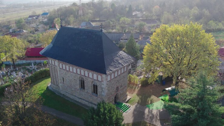 Aerial shot slowly pulls back from a historic medieval stone church, revealing a bright yellow autumn tree, a cemetery, and the surrounding rural village in Romania. Stefan the Great, Borzesti