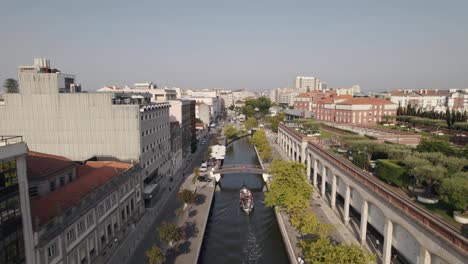 Aerial-descending-shot-of-moliceiro-boat-cruising-on-water-canal-under-pedestrian-bridge