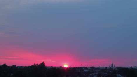 Lapso-De-Tiempo-De-La-Hermosa-Puesta-De-Sol-Roja-Escénica-Sobre-La-Ciudad-Con-El-Sol-Brillando-A-Través-De-Las-Nubes,-Plano-General