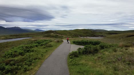 Aéreo-Siguiendo-Un-Longboard-Masculino-Despreocupado-A-Lo-Largo-De-Un-Camino-Vacío-En-El-Noroeste-De-Escocia