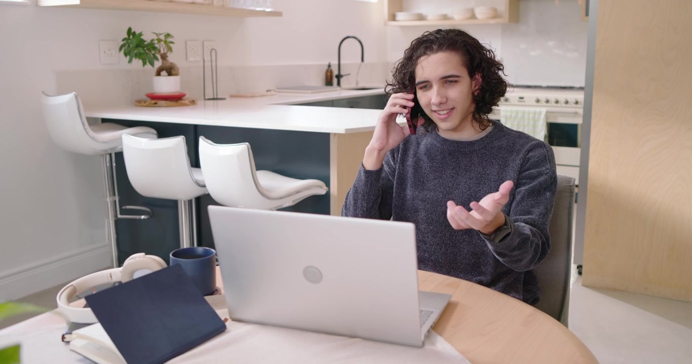 Afroamerikansk mand tager et telefonopkald i forbindelse med arbejde, gestikulerer, tapper på sin bærbare computer og smiler ved køkkenbordet.
