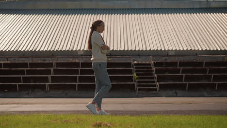 young woman in hoodie and jeans with long hair tied back walks near empty stadium seating on sunny day, arms folded, looking thoughtful, surrounded by empty bleachers and greenery