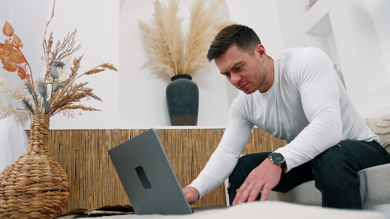 Frowning male leaned above his laptop on the coffee table. Man is typing on laptop trying to solve some issues. Low angle view.