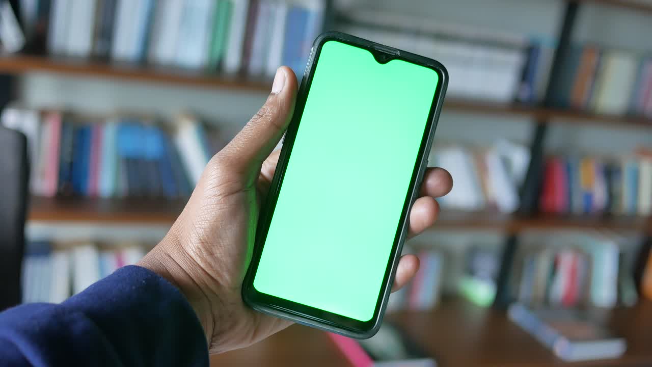 persona sosteniendo un teléfono inteligente con una pantalla verde en una biblioteca doméstica.
