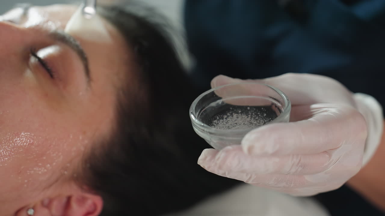 Close up of gloved hand holding small glass bowl of clear gel while brush picks up bubbles and applies smoothing strokes to client s cheek under bright spa light for deep hydration treatment