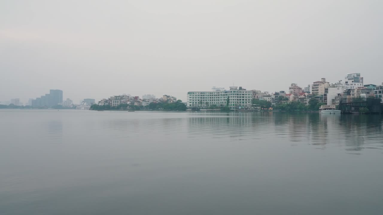 A rotating view over Hanoi’s West Lake in Vietnam, with calm waters, a misty skyline, and subtle reflections of buildings under a serene sky