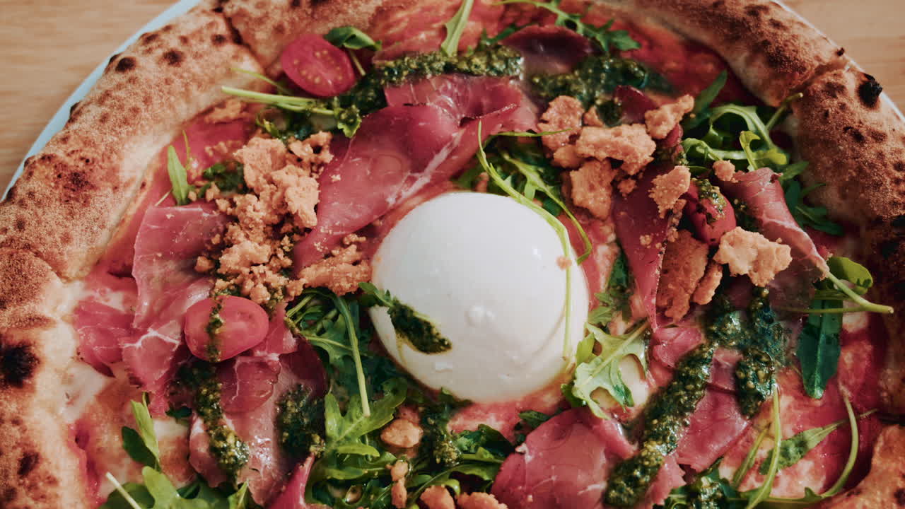 A beautifully shot overhead view of a gourmet pizza topped with burrata, prosciutto, cherry tomatoes, pesto, arugula, and crunchy crumbs