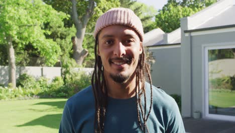 Portrait-of-biracial-man-with-hat-and-dreadlocks-outside-on-terrace-of-house-smiling-in-the-sun