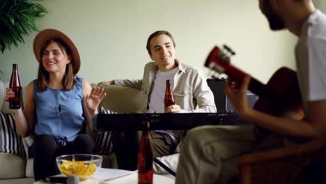 cheerful friends are having fun talking, singing and drinking beer during rehearsal. guitarist is playing, woman and man are clanging bottles and laughing.