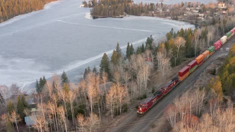 a long train carrying lots of shipping containers traveling through the forest beside a frozen lake in canada