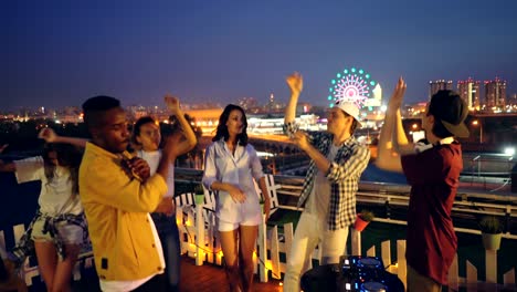 friends multiracial group are dancing on rooftop at night having open-air party while dj is working with equipment then clapping hands. partying, celebration and fun concept.