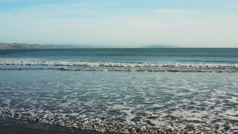 Waves-slowly-rolling-on-to-beach,-aerial-low-flyover-Bodega-Bay,-California,-USA