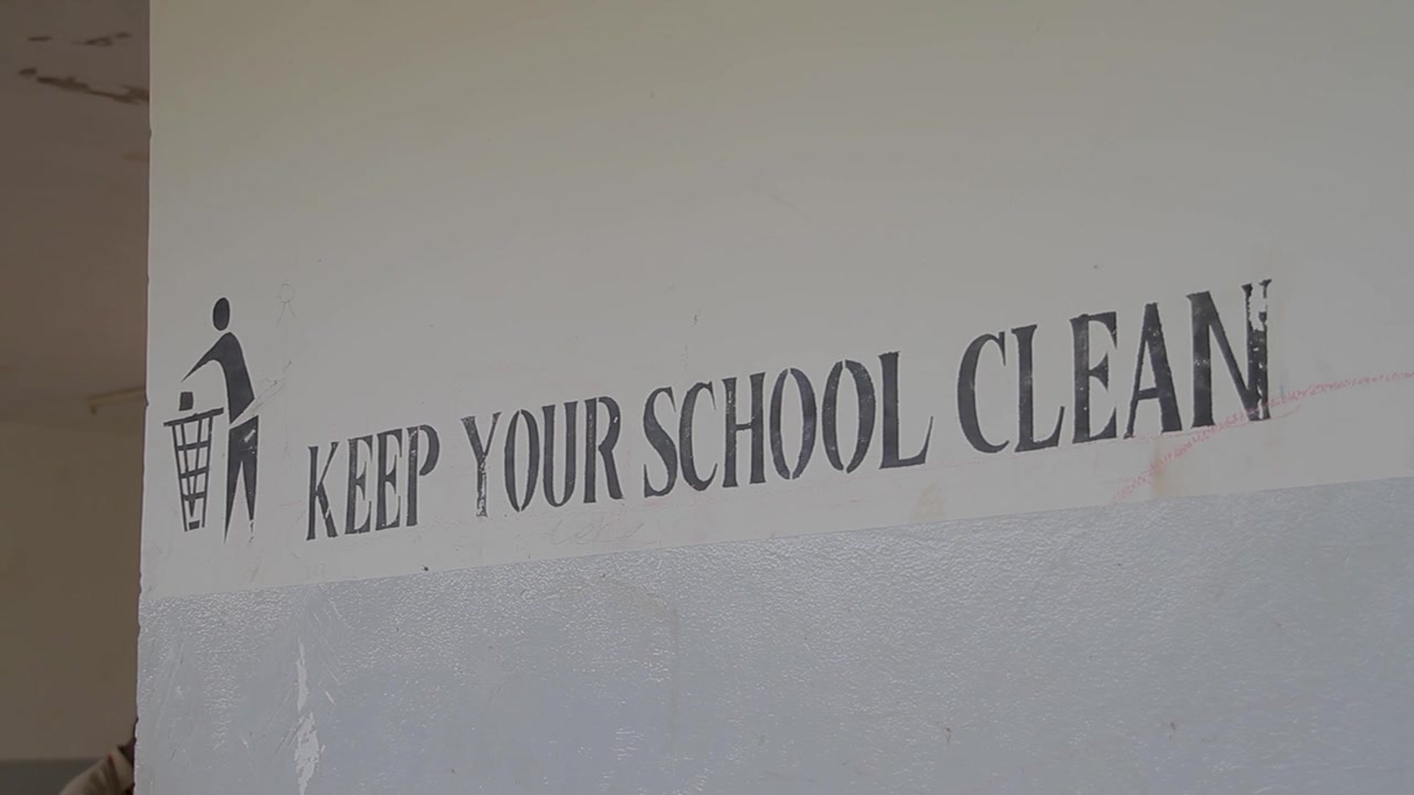 A close-up shot of a wall in a school building featuring a stencil-style sign that reads 'KEEP YOUR SCHOOL CLEAN' next to a trash can icon.
