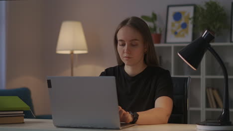 Una-Mujer-Joven-Y-Bonita-Está-Chateando-En-Línea-En-Las-Redes-Sociales-Por-La-Noche-Enviando-Mensajes-Por-Internet-En-Un-Cuaderno-Usando-Una-Computadora-Portátil-En-El-Retrato-Medio-De-La-Sala-De-Estar.